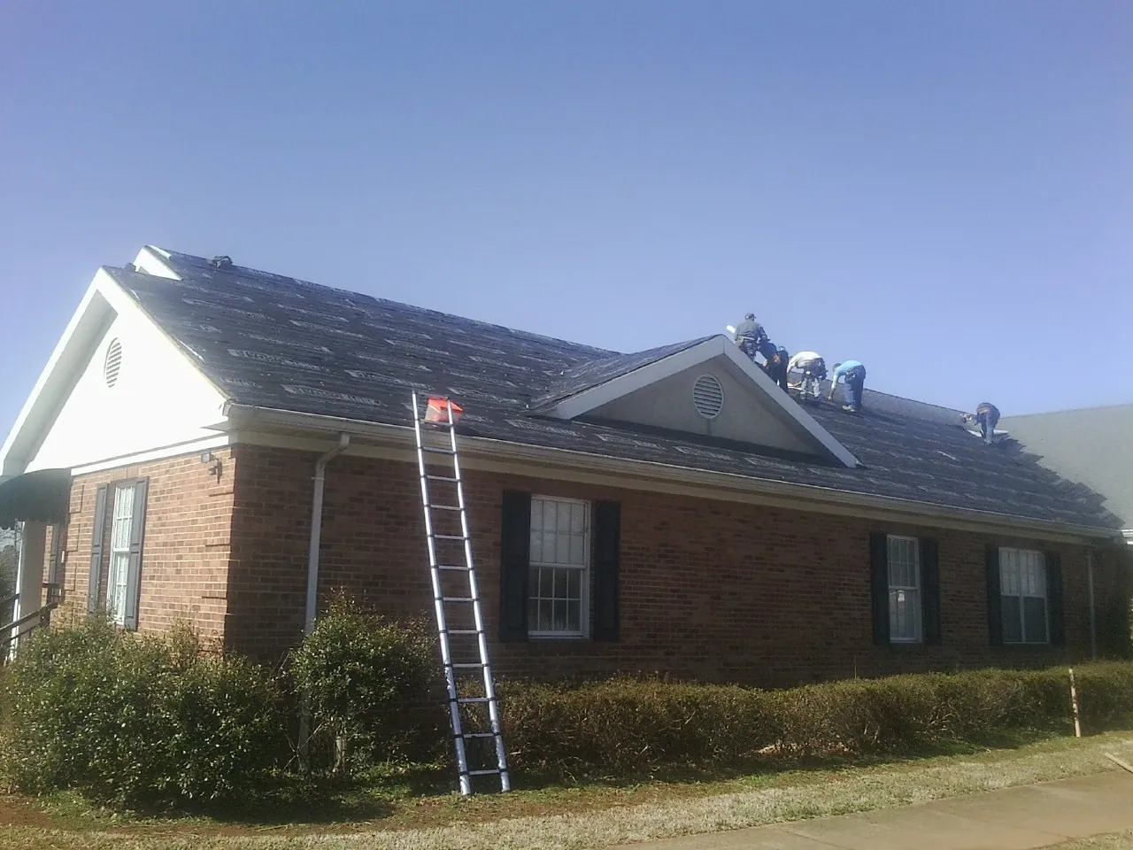 Crew tearing shingles off a brick ranch roof with a ladder set — Emergency Roof Repair work by our crew in Roaring River