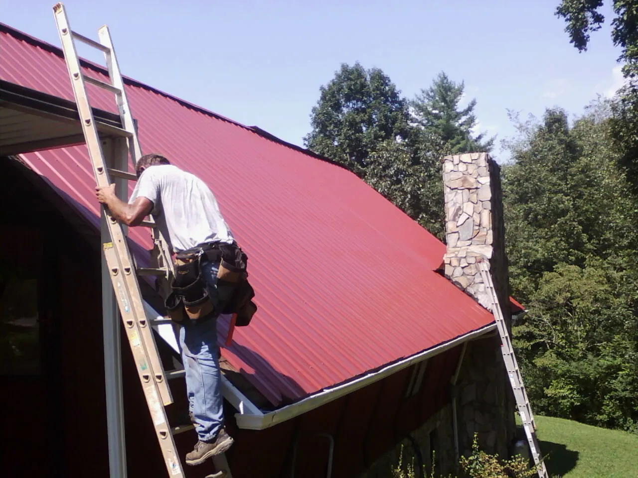 Roofer on a ladder beside a red metal roof and stone chimney — Soffit & Fascia Repair by Roofing Brothers in Roaring River